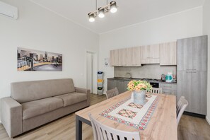 A modern kitchen with light wood cabinets, a dining table with a floral runner, and a sofa in a neutral color.