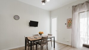Spiseområde med bord, stole, et fjernsyn ophængt på væggen og et vindue med gardiner.