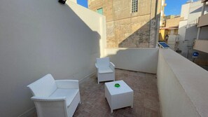 A small outdoor balcony with two white wicker chairs and a small table, surrounded by buildings.