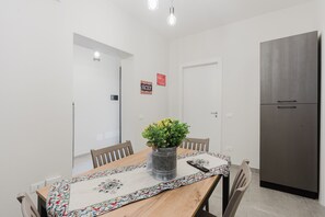 En matplats med ett träbord, stolar, en vas med blommor, ett mönstrat bordsduk, en fjärrkontroll och ett kylskåp.