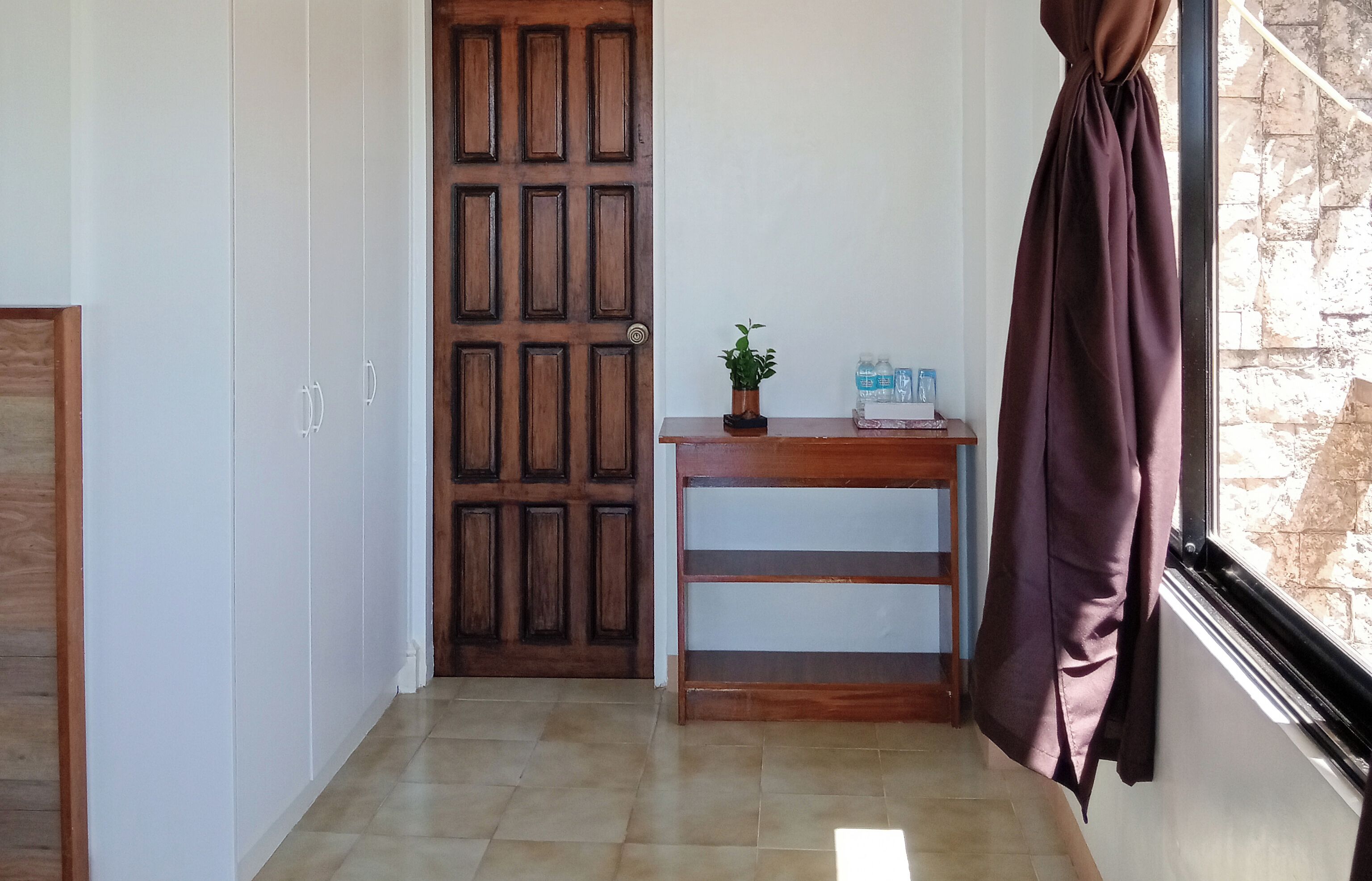 A hallway with a wooden door, a small table with a plant and water bottles, and a window with a curtain.