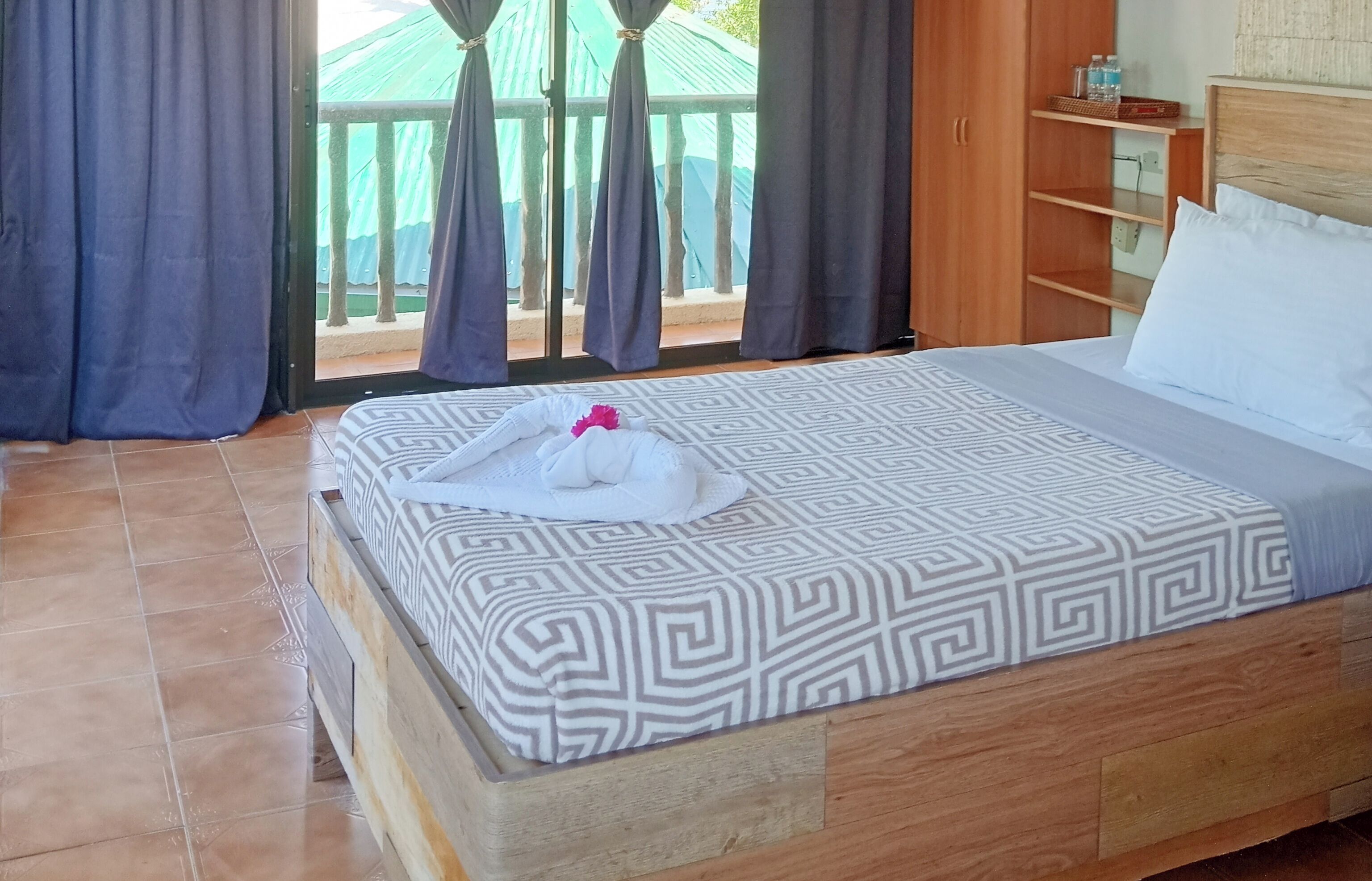 A neatly made bed with a patterned bedspread, a wooden headboard, and a view of a balcony with curtains.