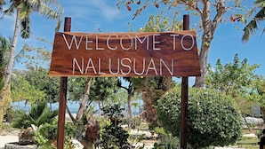 Een houten bord met de tekst 'WELCOME TO NALUSUAN', omringd door tropische begroeiing en palmbomen.
