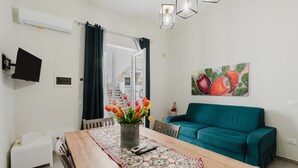 A living room with a wooden dining table, teal sofa, and a painting of fruits. - Apartment 'Lazzara 19 - Home 01' with Balcony, Wi-Fi and Air Conditioning (Marsala)