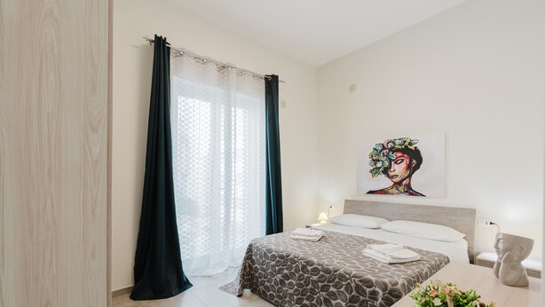 A bedroom with a bed, a bedside table, a lamp, a wall art piece, and a window with curtains. - Apartment 'Lazzara 19 - Home 01' with Balcony, Wi-Fi and Air Conditioning (Marsala)