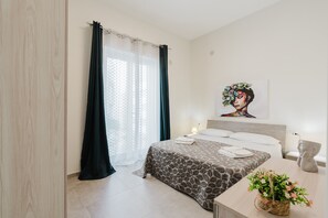 A bedroom with a bed, a bedside table, a lamp, a wall art piece, and a window with curtains. - Apartment 'Lazzara 19 - Home 01' with Balcony, Wi-Fi and Air Conditioning (Marsala)