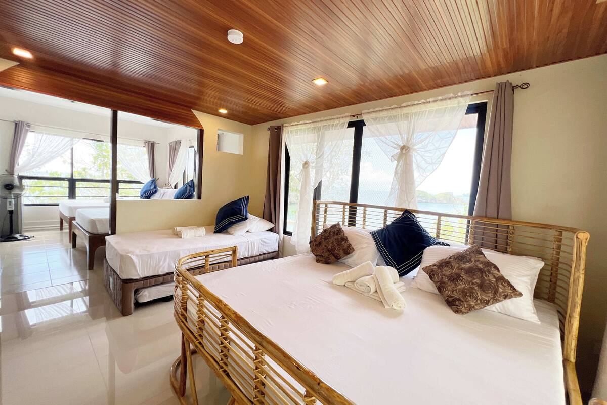 Ein Hotelzimmer mit Holzboden, zwei Betten mit weißen Leinentüchern, einem Holzstuhl und großen Fenstern mit transparenten Vorhängen.