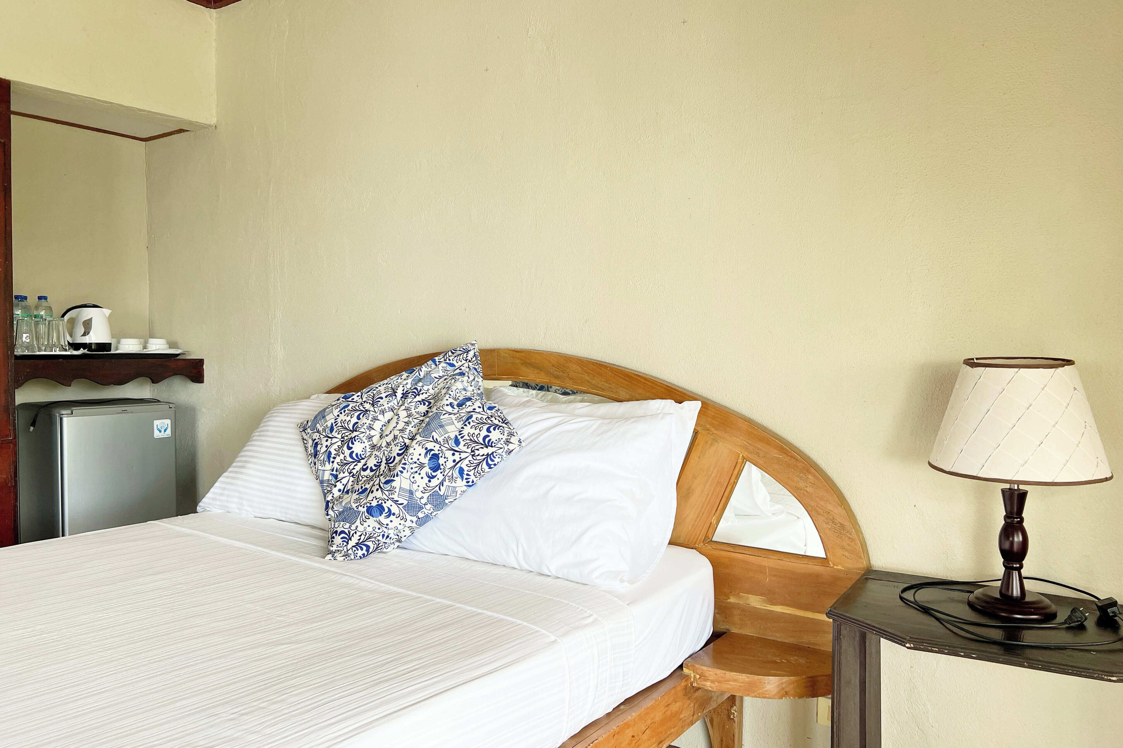 Un letto ben rifatto con una testiera in legno, un comodino con una lampada e un piccolo frigorifero sul lato sinistro.