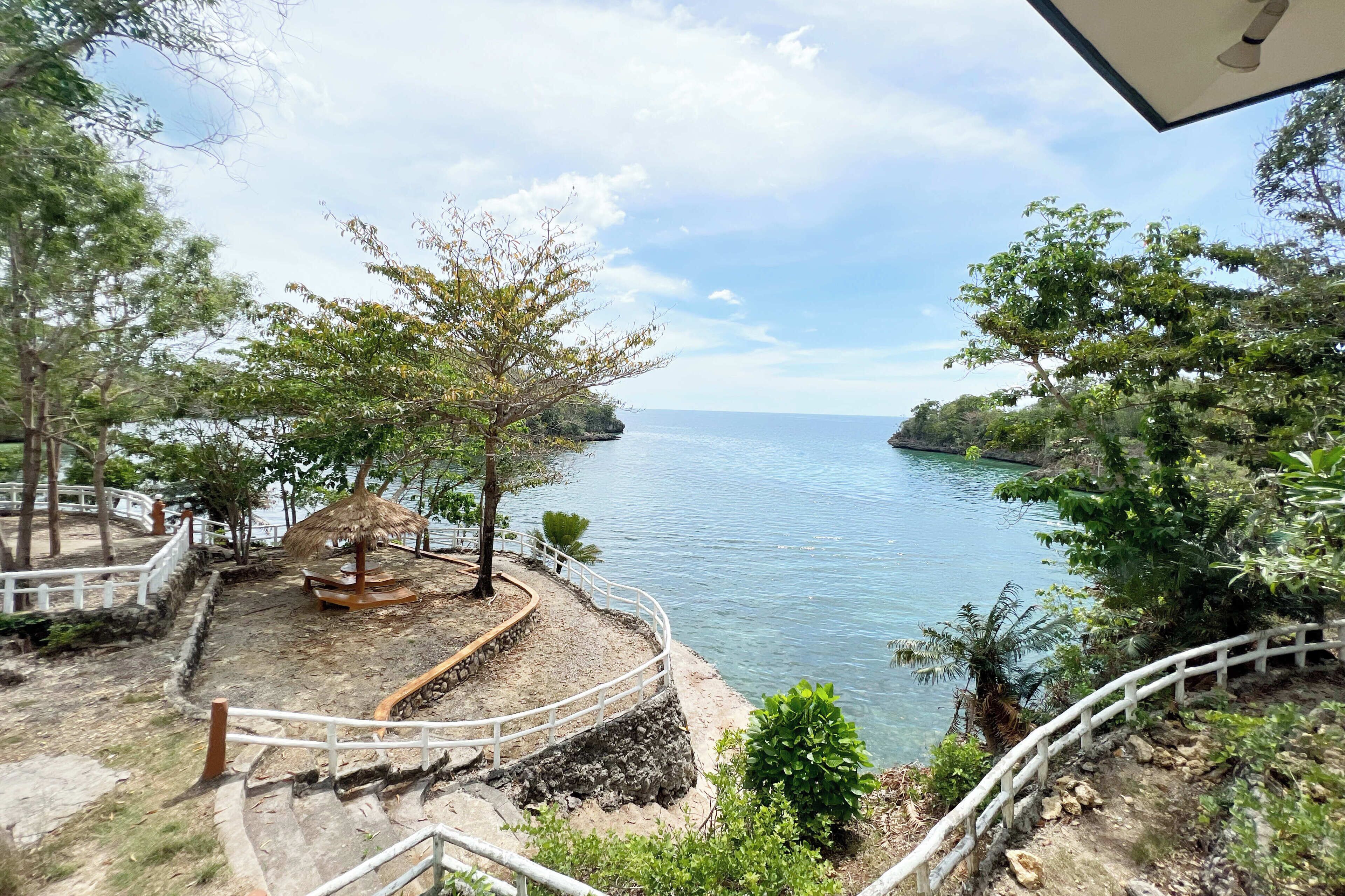A coastal walkway with a thatched structure, surrounded by trees and a railing.