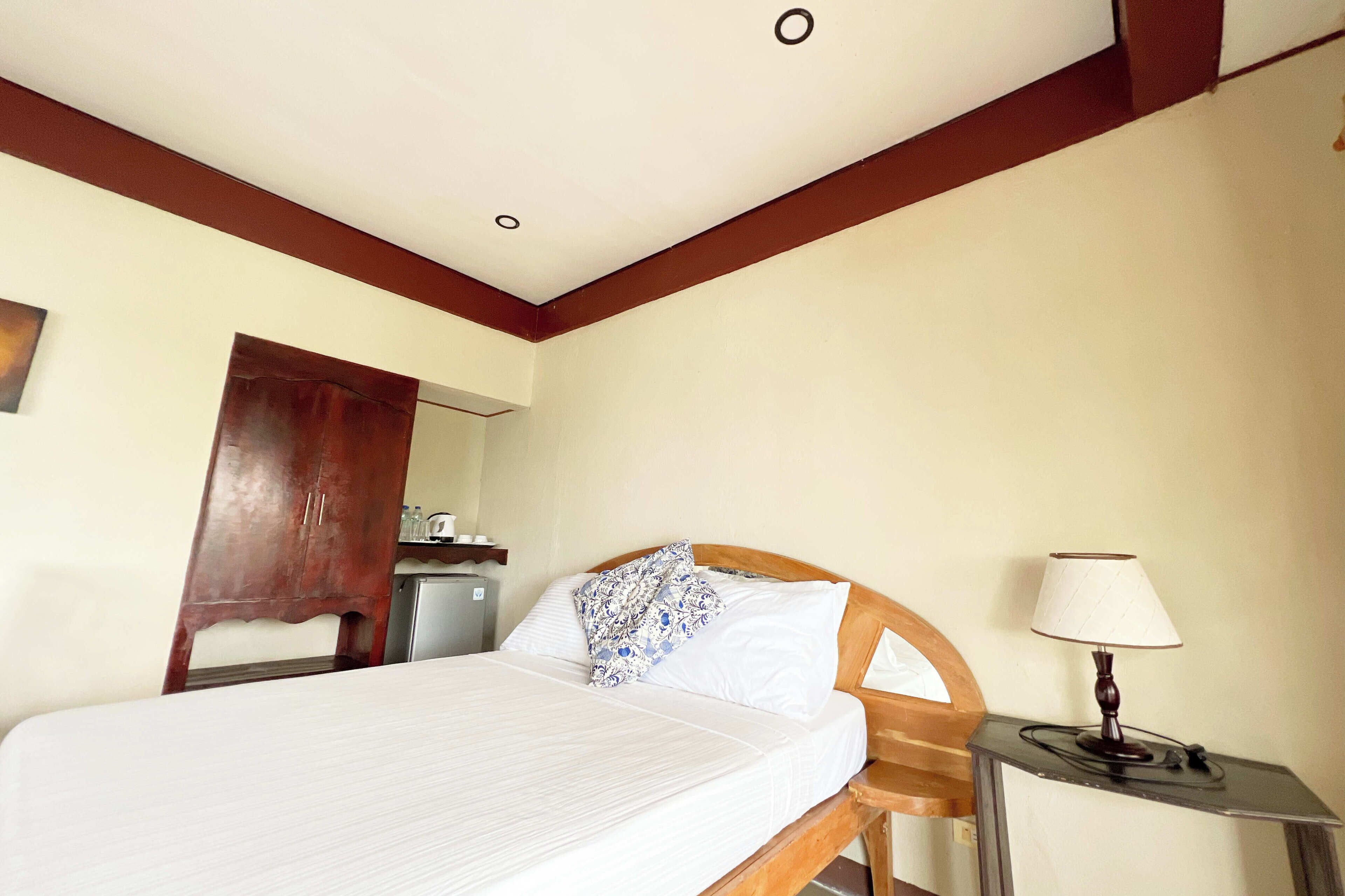 Habitación de hotel con cama, cabecero de madera, mesita de noche con lámpara y un escritorio pequeño con lámpara.