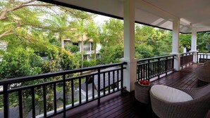 Un espace extérieur abrité, doté d'une terrasse en bois, de meubles en osier et offrant une vue sur une végétation luxuriante et des bâtiments en arrière-plan.