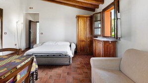 Un dormitorio con cama, un armario de madera, una ventana con persianas, una silla y una mesa con mantel estampado.