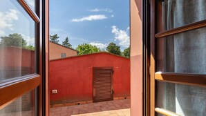 Visão pela janela de um edifício vermelho com porta marrom, pátio com azulejos e céu claro.