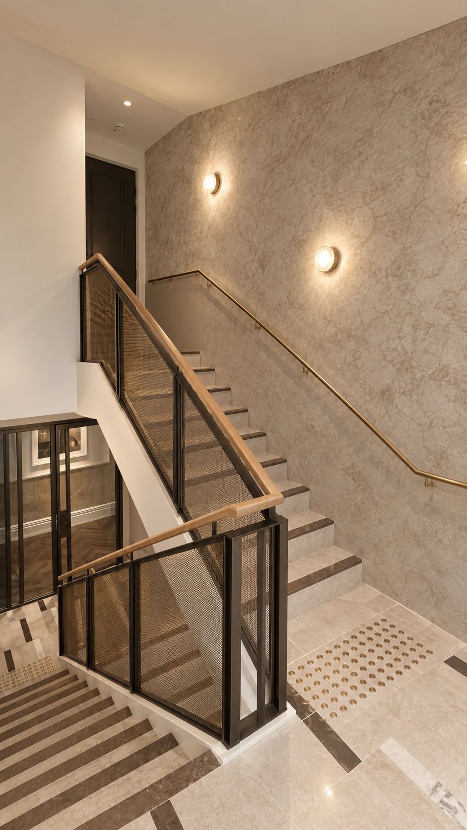A modern staircase with glass railings and a wooden handrail, leading to a room with a herringbone-patterned floor.