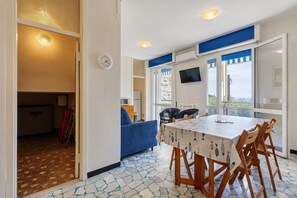 A dining area with a table and chairs, a sofa, a TV, and a view through the windows.