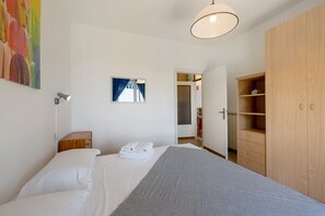 A bedroom with a bed, a bedside table with towels, a wardrobe, a painting, and a window with curtains.