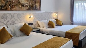 A hotel room with two beds, each with white linens and mustard-colored throw pillows. There is a large abstract painting above the beds, and a wall-mounted light fixture on the headboard.