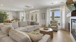Un salon spacieux avec un grand canapé d’angle, un canapé plus petit et une table basse. On y trouve une cheminée, un coin repas avec des meubles en osier, et de grandes fenêtres qui offrent une vue sur les arbres et les montagnes.