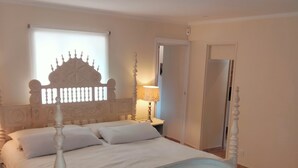 A bedroom with a four-poster bed, a white upholstered chair, a patterned rug, and a nightstand with a lamp. - Clos saint Joseph (Orange)