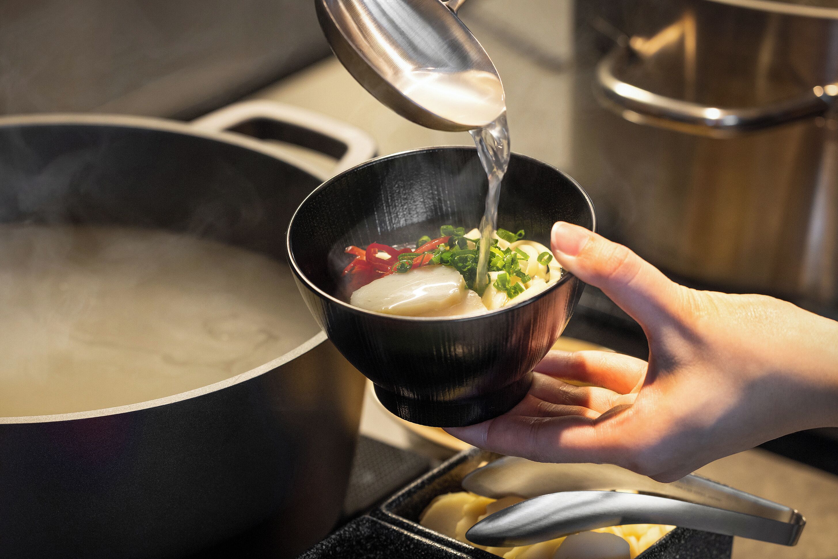 A hand holding a bowl of soup with a lid being lifted, revealing steam.