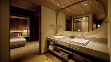A modern hotel bathroom with a double sink, a large mirror, and a bed in the adjacent room.
