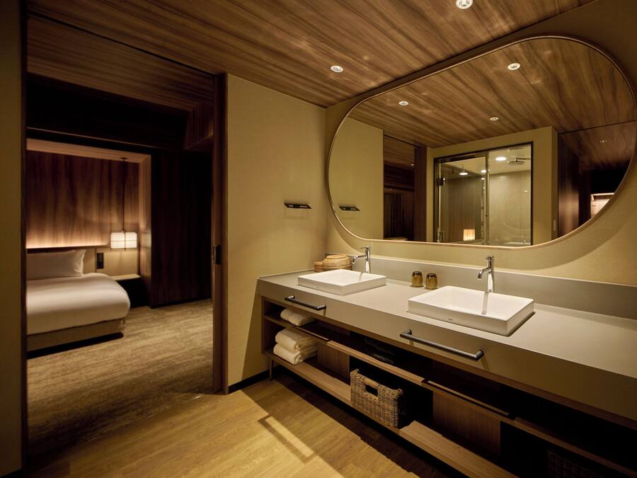 A modern hotel bathroom with a double sink, a large mirror, and a bed in the adjacent room.