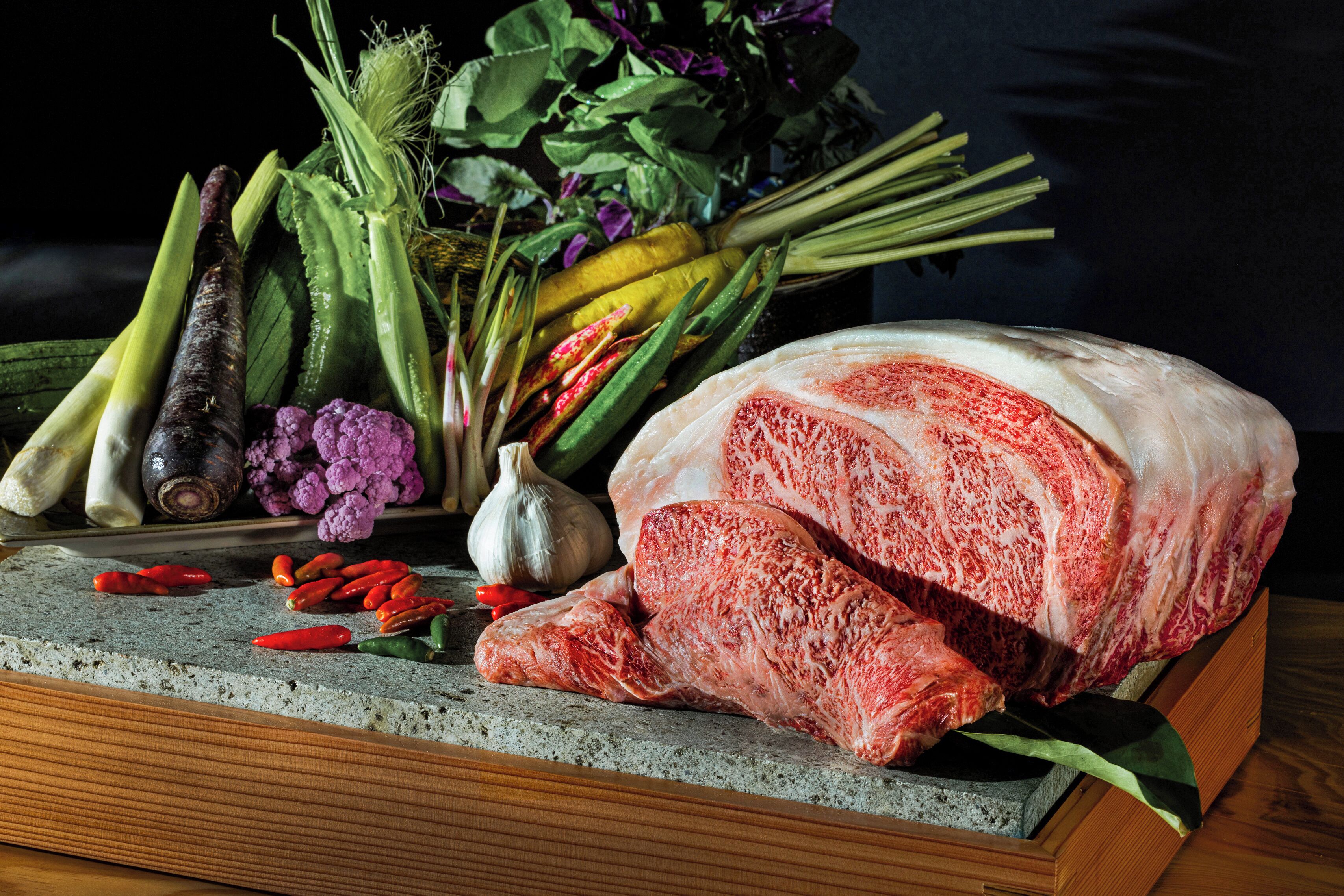 A variety of fresh vegetables and a large piece of meat on a stone surface.