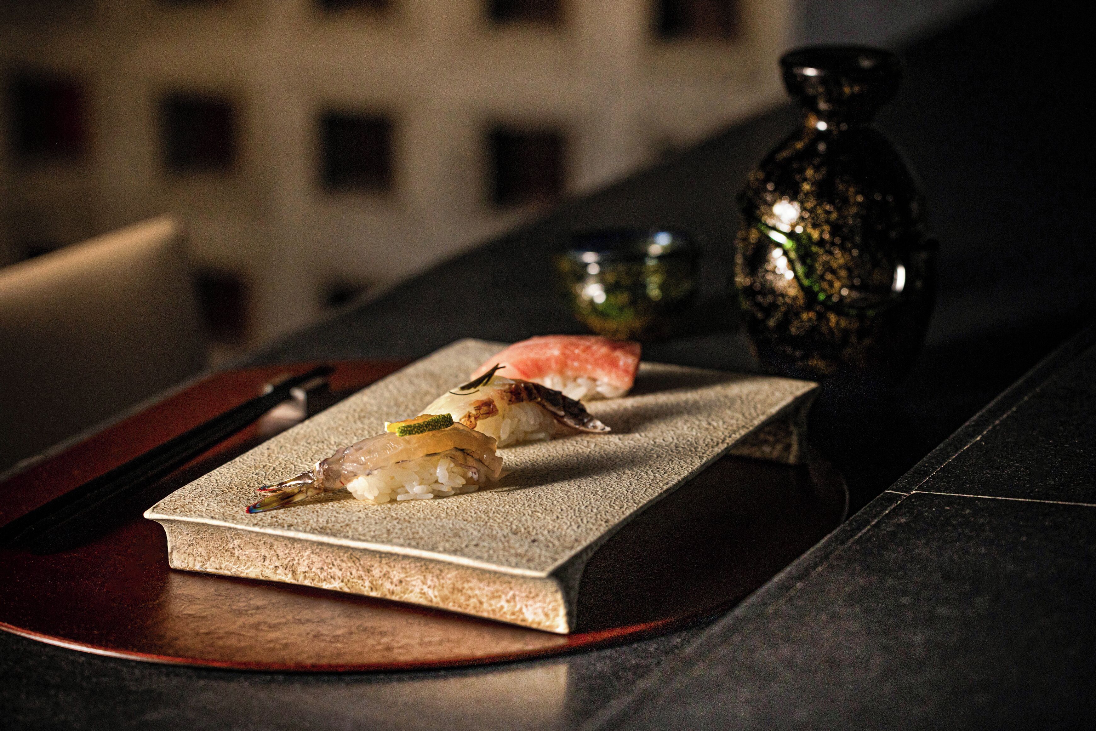 A sushi dish on a rectangular plate with chopsticks and a small bowl, set on a dark surface with a building in the background.