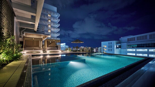 Un espace piscine nocturne avec un bâtiment moderne, un espace salon couvert et une vue sur les gratte-ciel de la ville.