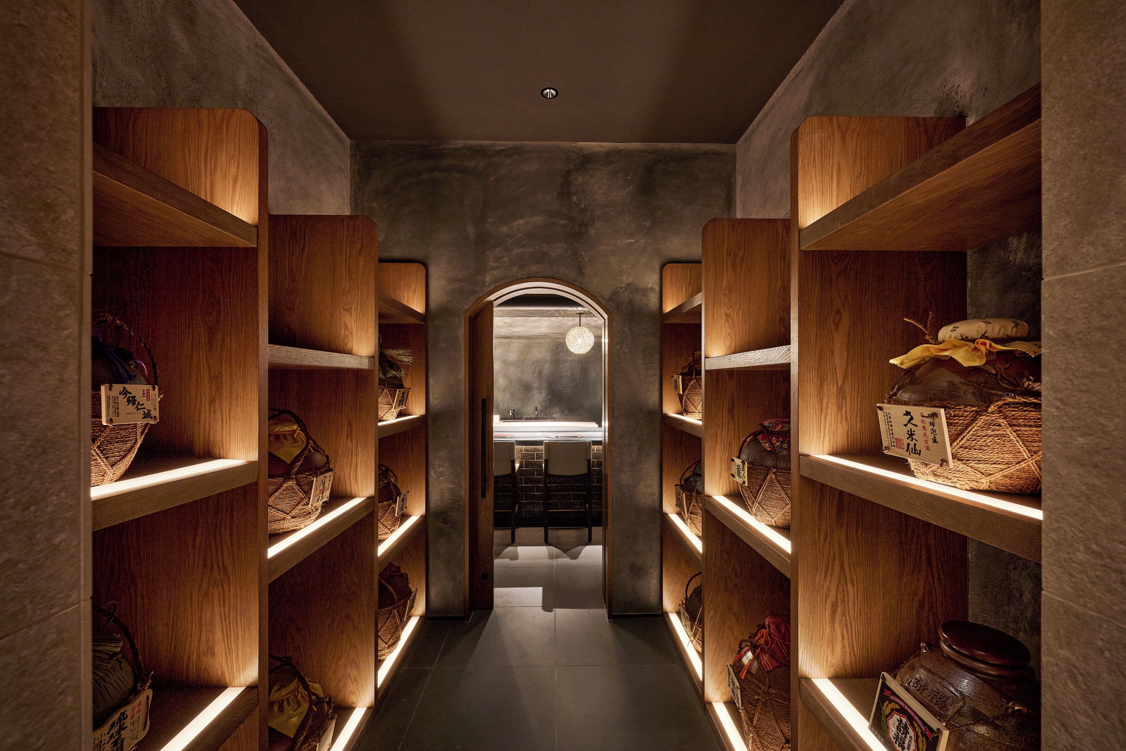 Un couloir étroit, avec des étagères en bois de chaque côté, qui mène à une salle de bain équipée d’un lavabo et d’un miroir.