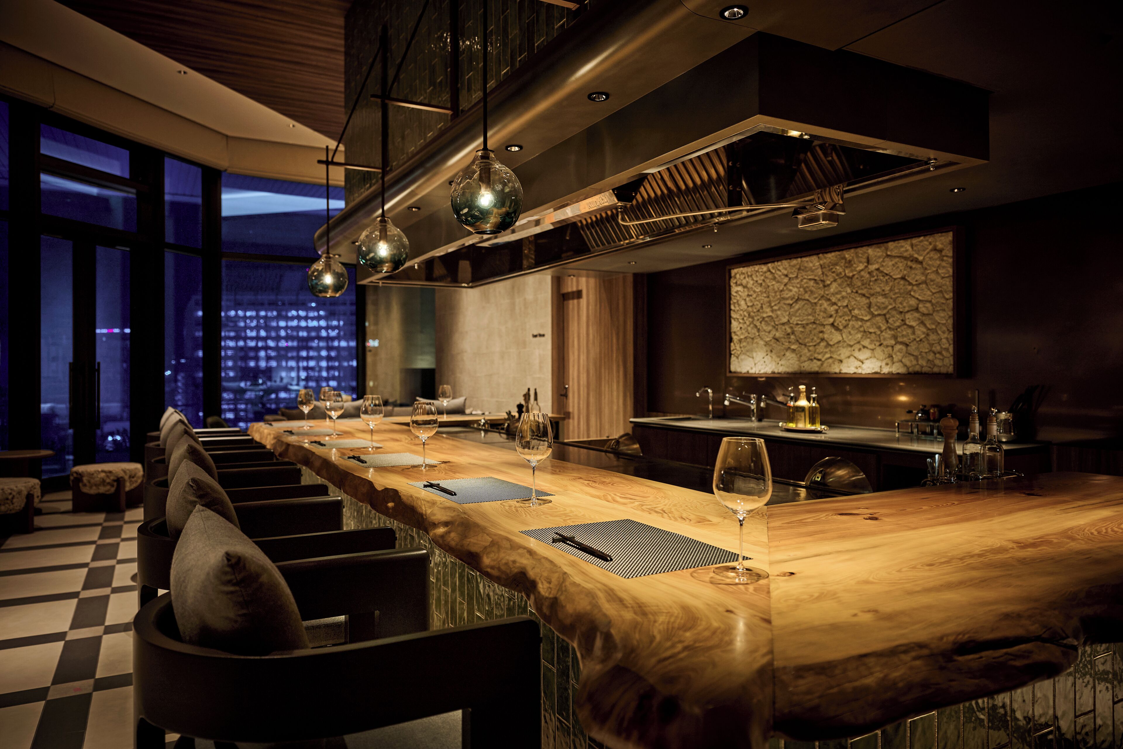 A modern bar area with a long wooden counter, seating with cushions, and a city view through large windows.