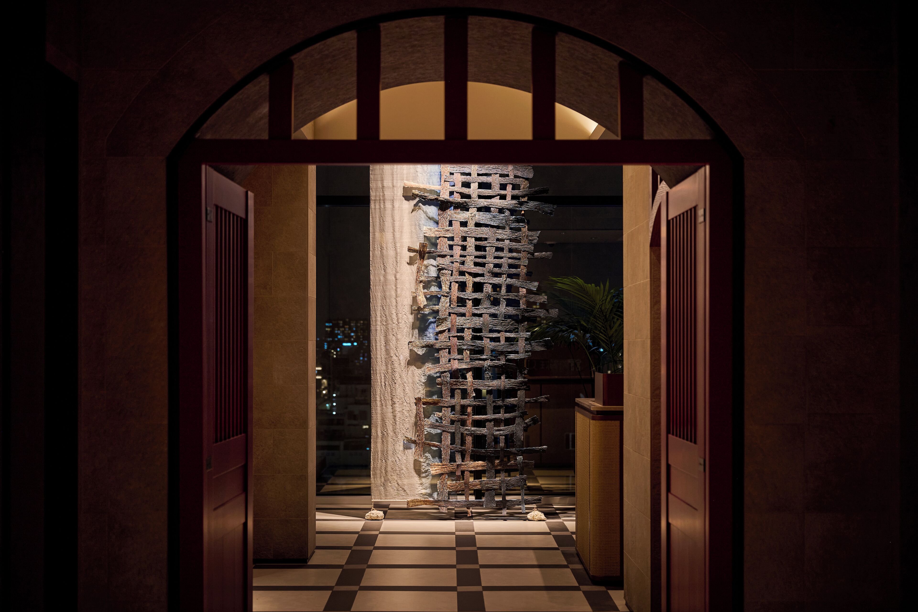 A hallway with a checkered floor, a wooden column with a lattice structure, and a potted plant.
