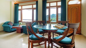 Een eetgedeelte met een ronde glazen tafel, omringd door houten stoelen met blauwgroene kussens, een bank en een bijzettafeltje.