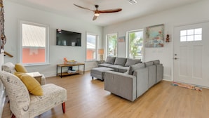 Un salon avec un canapé d’angle gris, un fauteuil à motifs, un ventilateur de plafond et un téléviseur à écran plat.