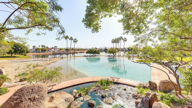 A large outdoor swimming pool surrounded by rocks and trees, with a view of a distant building. - Val Vista Lakes Extended Getaway: Hot Tub Access! (Gilbert)