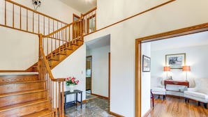 A wooden staircase leading to an upper floor, a tiled floor, a small table with a vase of flowers, and a seating area with two lamps and a framed picture on the wall.