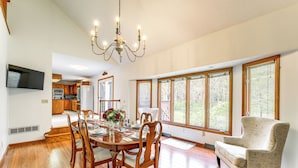 A dining room with a wooden table, chairs, and a chandelier. There is a kitchen in the background with wooden cabinets and a refrigerator.