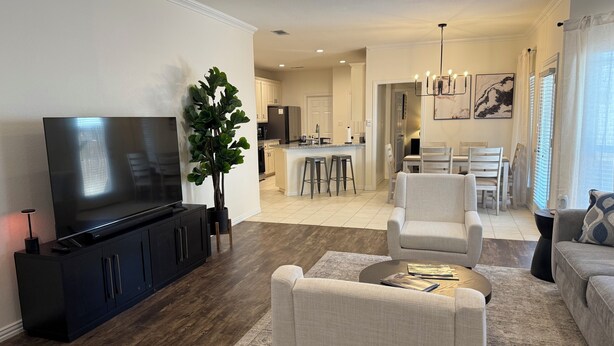 A modern living room with a flat-screen TV, a large potted plant, and a dining area with a kitchen in the background.
