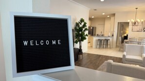 A modern living room with a 'WELCOME' sign on a wall-mounted panel, a dining area with a kitchen in the background, and a large potted plant.