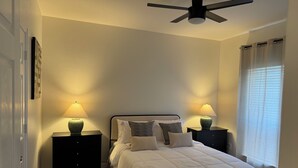 A bedroom with a bed, two bedside tables with lamps, a ceiling fan, and a window with curtains.