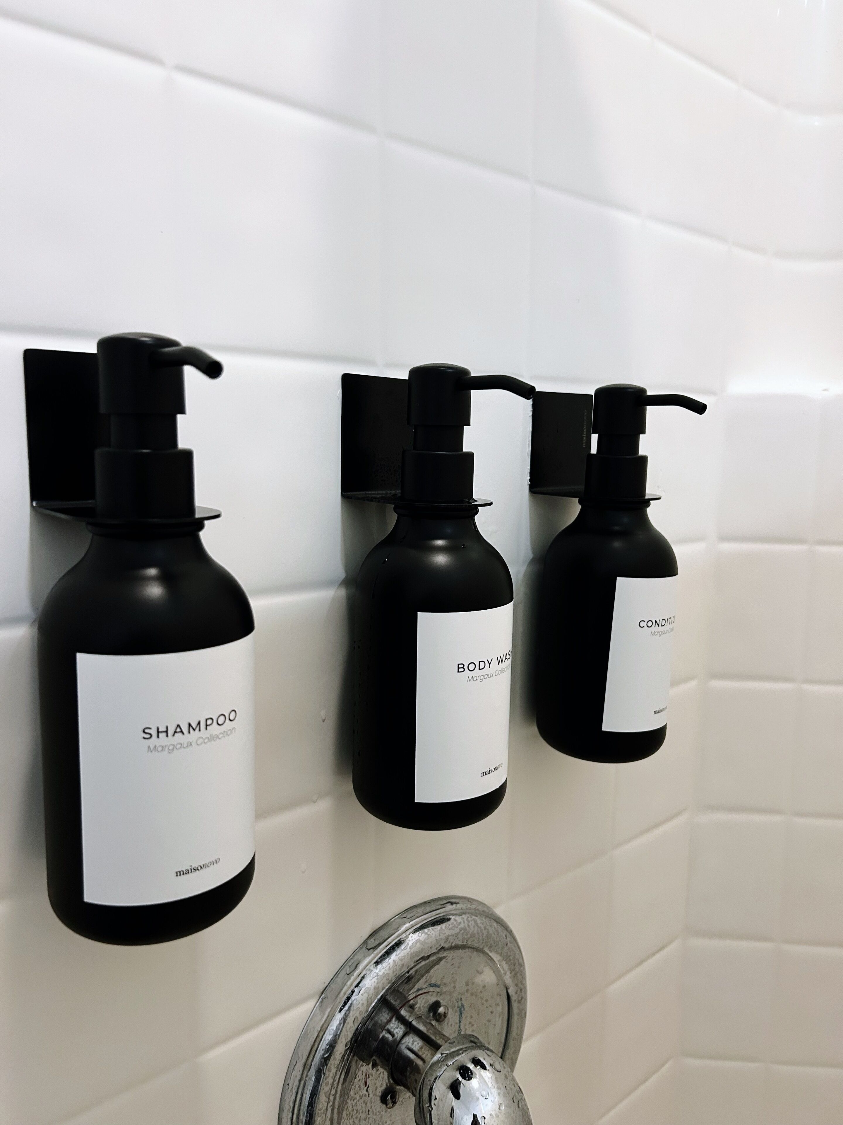 Three black bottles of shampoo and conditioner on a white tiled wall.