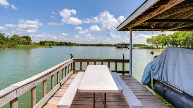 A wooden deck with a picnic table and benches overlooking a calm lake, with a covered area and a boat docked nearby. - Sunrise Cove RC LLC: Lakefront Home w/ 2 Casitas! (Kerens)
