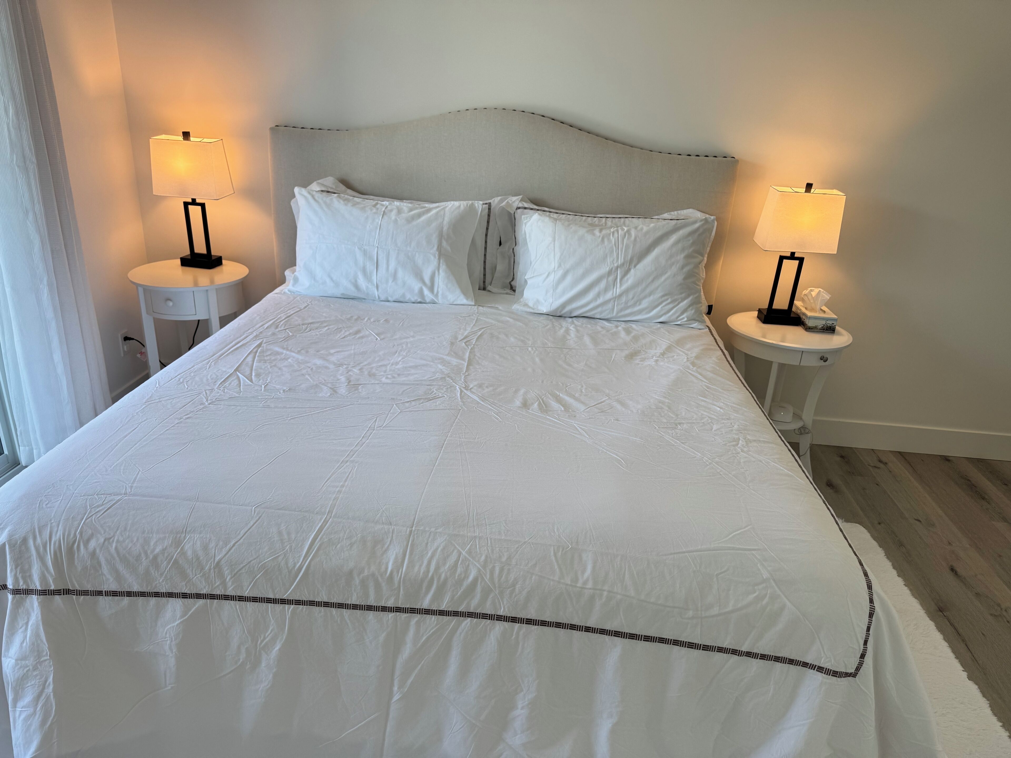 A neatly made bed with white linens and a decorative border, flanked by two bedside tables with lamps, in a room with a simple, clean design.