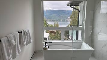A modern bathroom with a freestanding bathtub, a large window offering a view of a lake and mountains, and a towel rack with white towels.