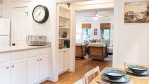 A kitchen with white cabinets, a clock, and a dining area with a table set for two. - The Long Time PET FRIENDLY Cape Fear Station (bald head island)