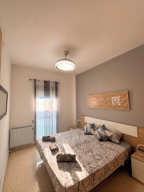A bedroom with a bed, a bedside table, a nightstand, a ceiling fan, a radiator, and a window with curtains.