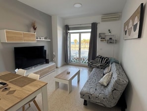 A modern living room with a flat-screen TV, a wooden dining table, a sofa with a patterned cover, and a balcony with a view. - FIRST LINE BEACH PARTMENT (València)