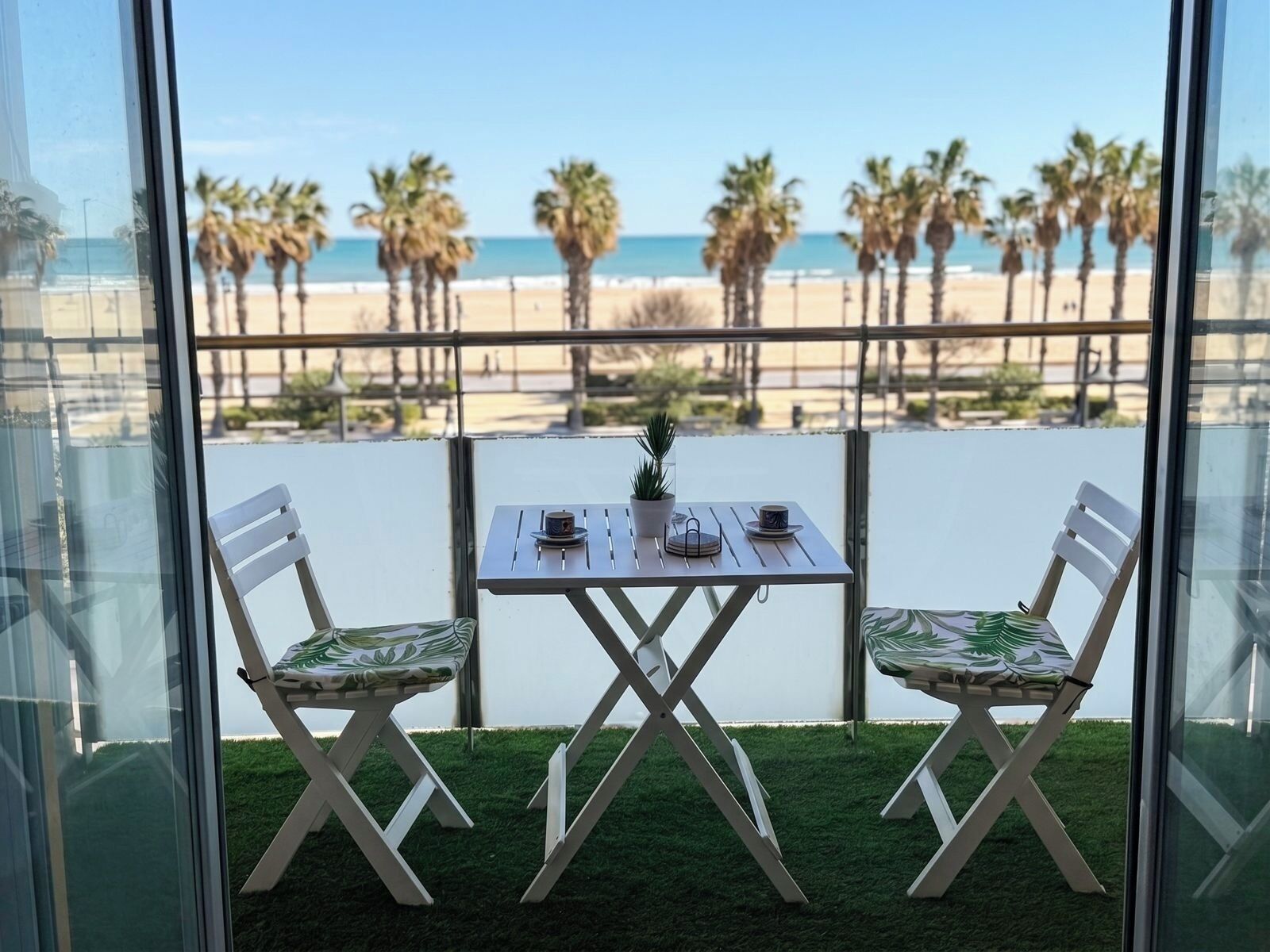 A balcony with a table and two chairs, overlooking a beach with palm trees and the ocean.