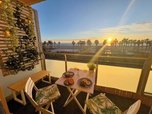 A balcony with a table set for two, overlooking a beach with palm trees and the ocean.