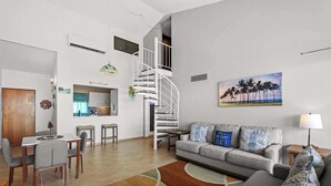 Un salon moderne avec un canapé gris, une table à manger avec des chaises, un escalier en colimaçon et un tableau sur le thème de la plage accroché au mur.